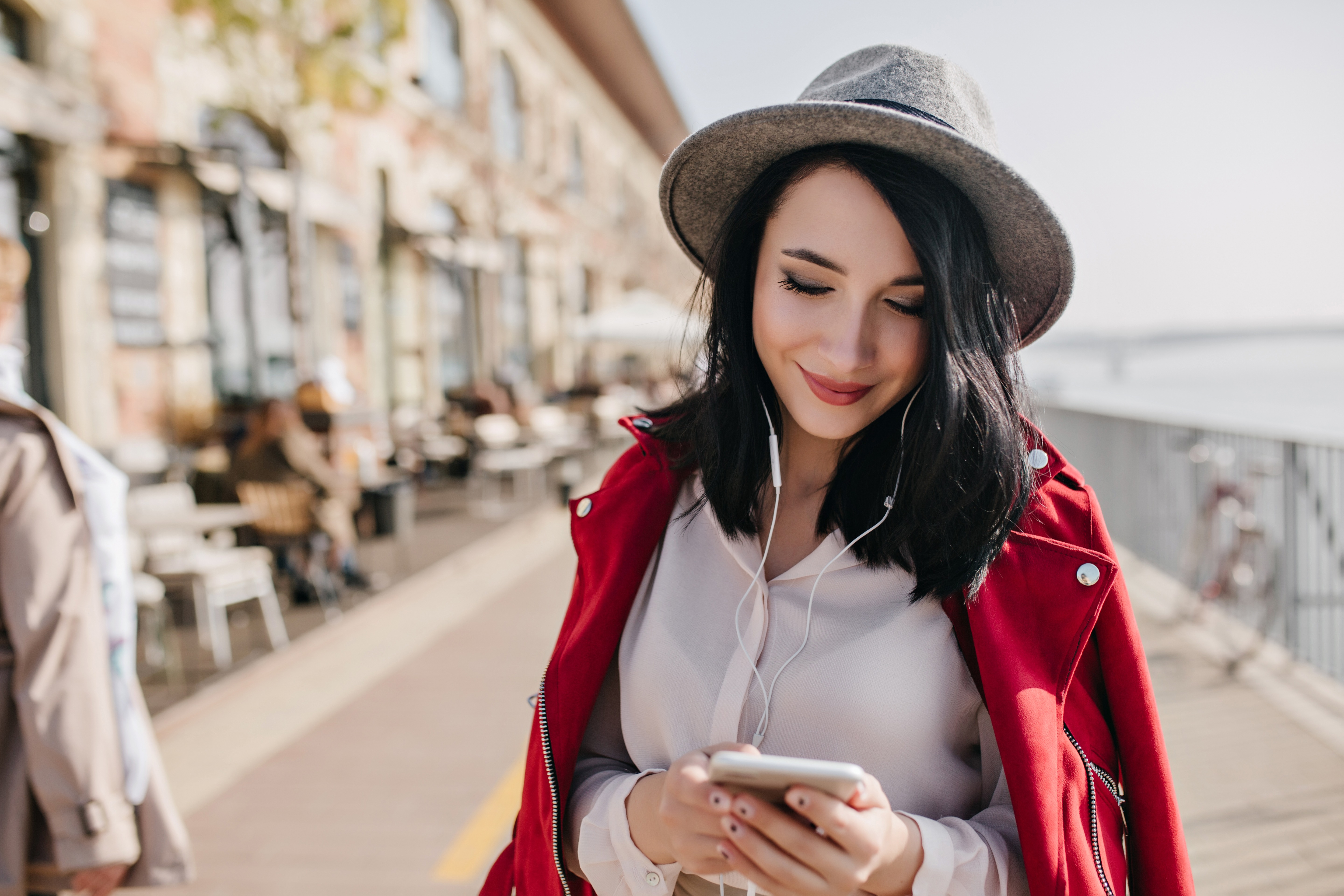Charming black-haired girl in earpones walking around city and listening music. Lovely woman in gray hat texting message on smartphone on blur urban background..
