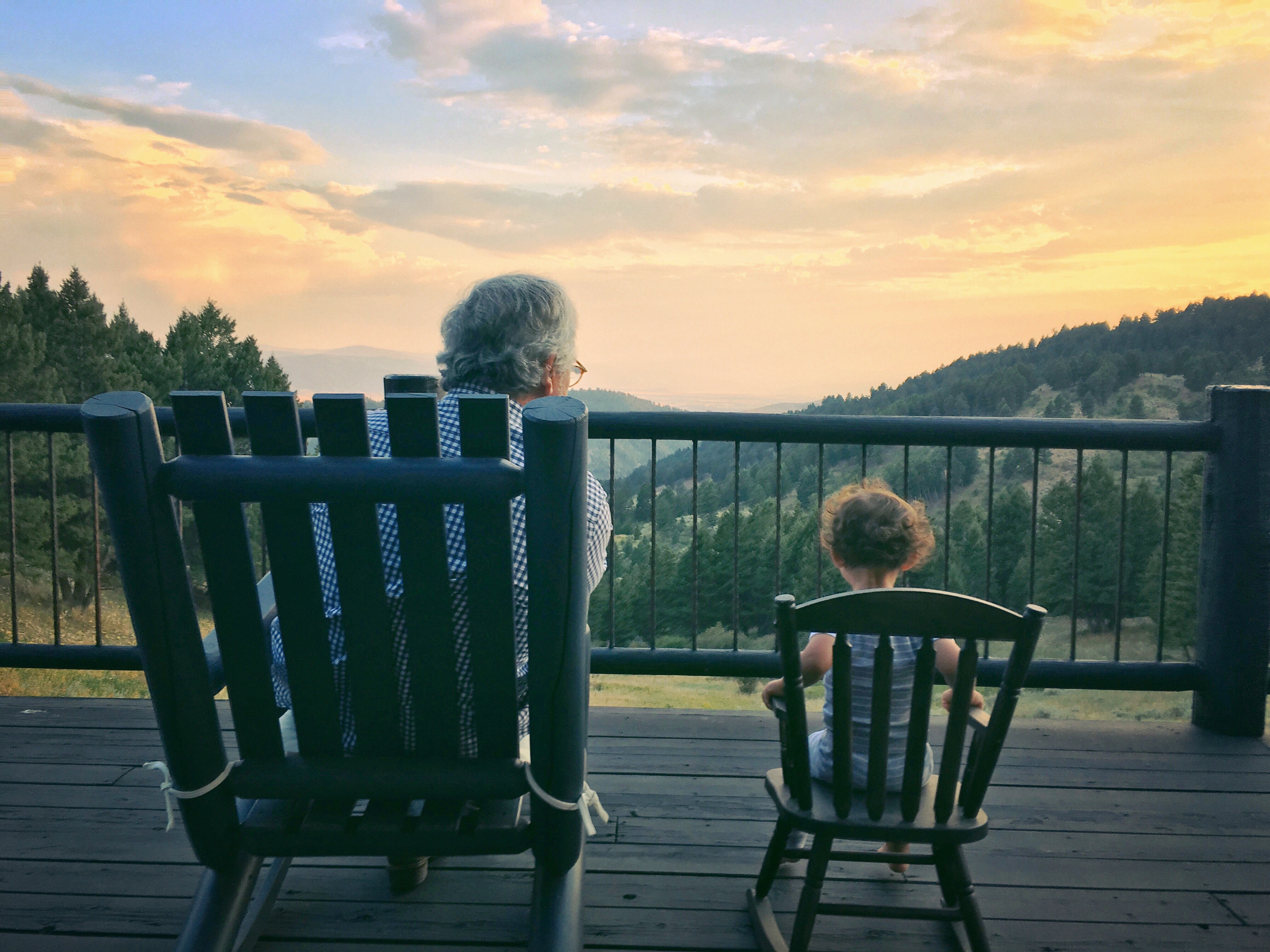 Grandfather With Granddaughter Sitting On Chair At Observation Point Against Cloudy Sky During Sunset