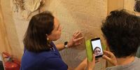 A blind woman kneels in front of a wall in the museum to touch the writing engraved in its plasterwork. This happens as a second woman looks at someone on her cellphone who is observing this exploration via WhatsApp. (Photo: Craig Strachan)