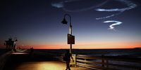 A person walks on a pier beneath the contrail remaining from a SpaceX Falcon 9 rocket carrying a payload of 22 Starlink internet satellites into space after launching from Vandenberg Space Force Base on 1 April 2024 in San Clemente, California.  (Photo:  Mario Tama / Getty Images)