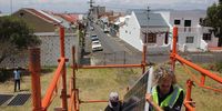 A member of the SOLA Future Energy team and YouLead Warrior mentor, Kutlwano Nondabula, carry a solar module up the scaffolding stairs onto the roof of a classroom at Salt River Secondary School. In the background are the suburbs of Salt River. Photo: Christi Nortier