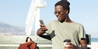 Outdoor portrait of fashionable smiling African American male tourist enjoying coffee out of paper cup and searching for new interesting places on websites using online application on mobile phone