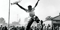 Hugh Masekela leaps into the air with delight after receiving a trumpet from<br>American jazz legend, Louis Armstrong, 1956. Hugh Masekela would go on to become a<br>legendary South African jazz musician, but in this image, he was still a teenager, and the<br>trumpet came to him via Trevor Huddleston, an anti-apartheid activist and priest. Almost half a<br>century later, the image was chosen for the cover of a book on Masekela&#39;s life, Sophiatown,<br>Johannesburg, © Alf Kumalo Family Trust]