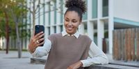 Happy beautiful woman with curly hair gathered in bun records selfie video or poses for photo uses smartphone dressed in neat clothes poses outdoors in urban setting. Modern lifestyle concept